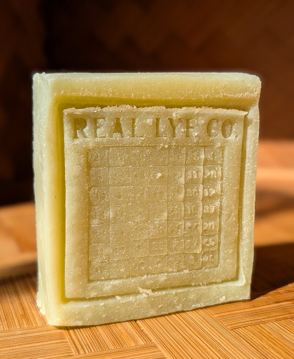 A square pale yellow bar of soap standing upright on a wooden surface, embossed with the brand name 'REAL LYE CO.' and a grid design.