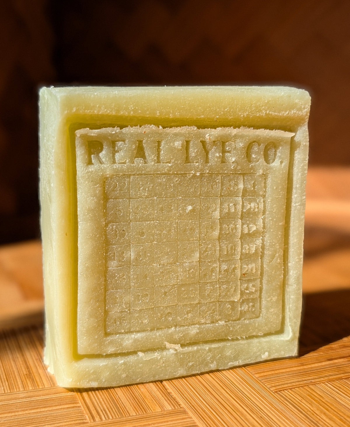 A square pale yellow bar of soap standing upright on a wooden surface, embossed with the brand name 'REAL LYE CO.' and a grid design.