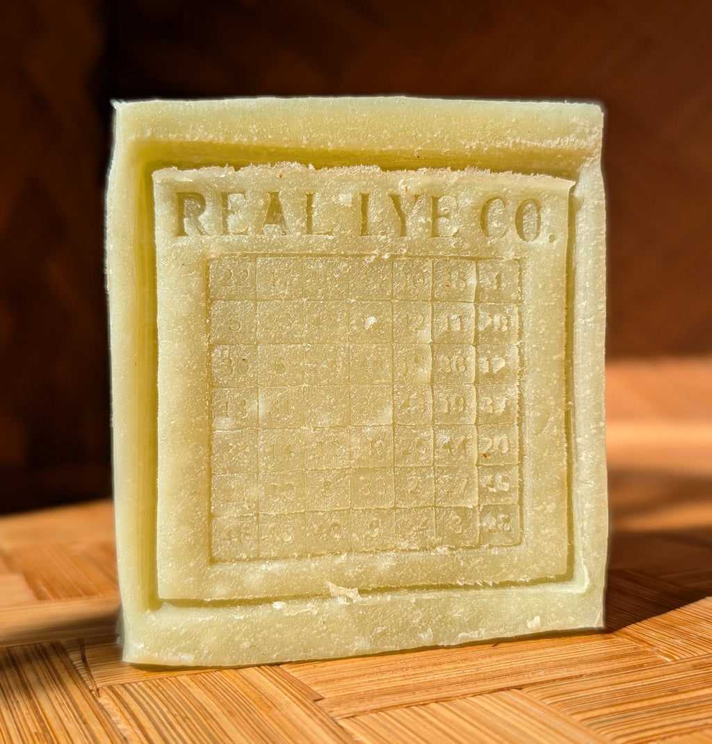 A square pale yellow soap bar embossed with 'REAL LYE CO.' and a grid design, standing on a wooden surface with a warm, dark wood background.