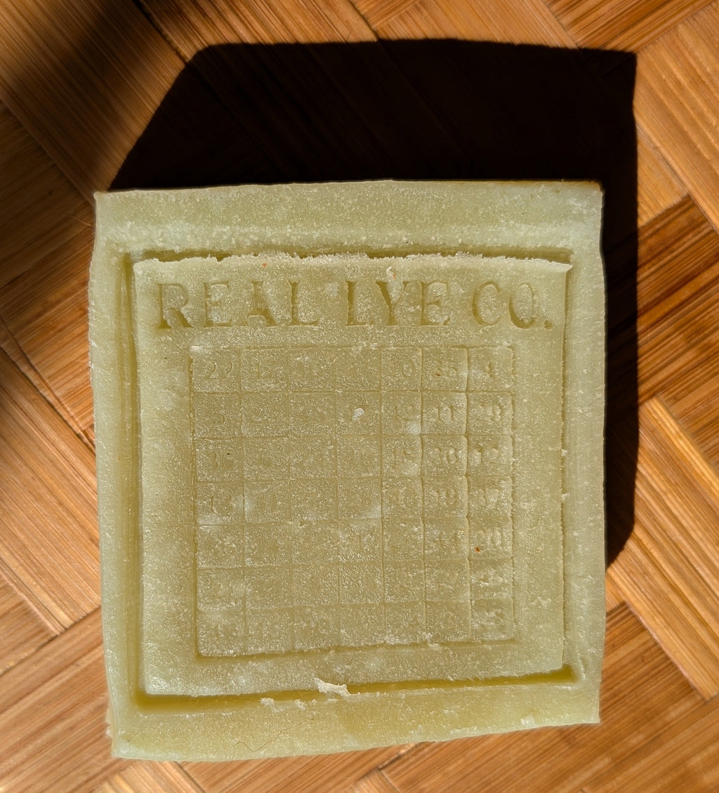 A rectangular bar of pale yellow-green soap with 'REAL LYE CO.' embossed on the top, placed on a brown woven wood surface.