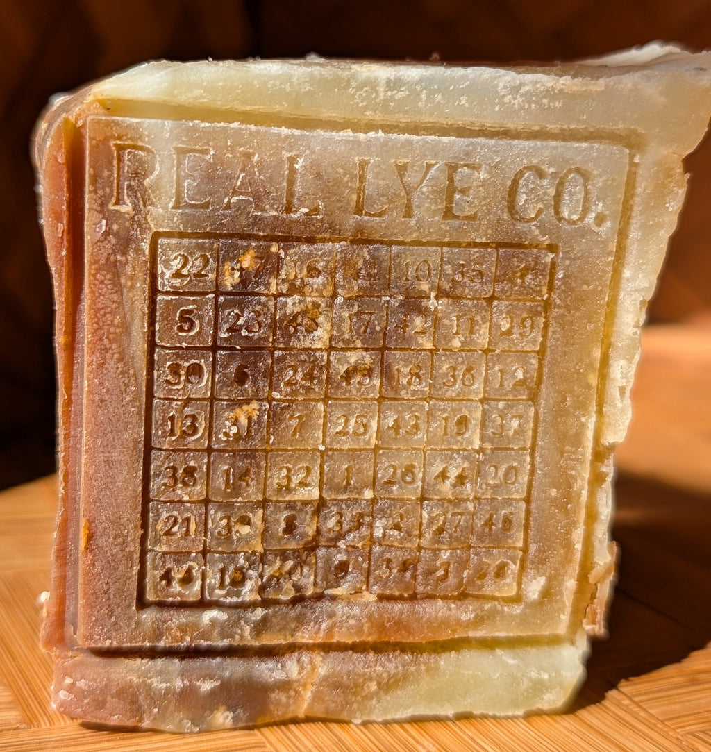 A rectangular bar of handmade soap embossed with 'REAL LYE CO.' and a numbered grid, resting on a wooden surface with a warm, rustic background.
