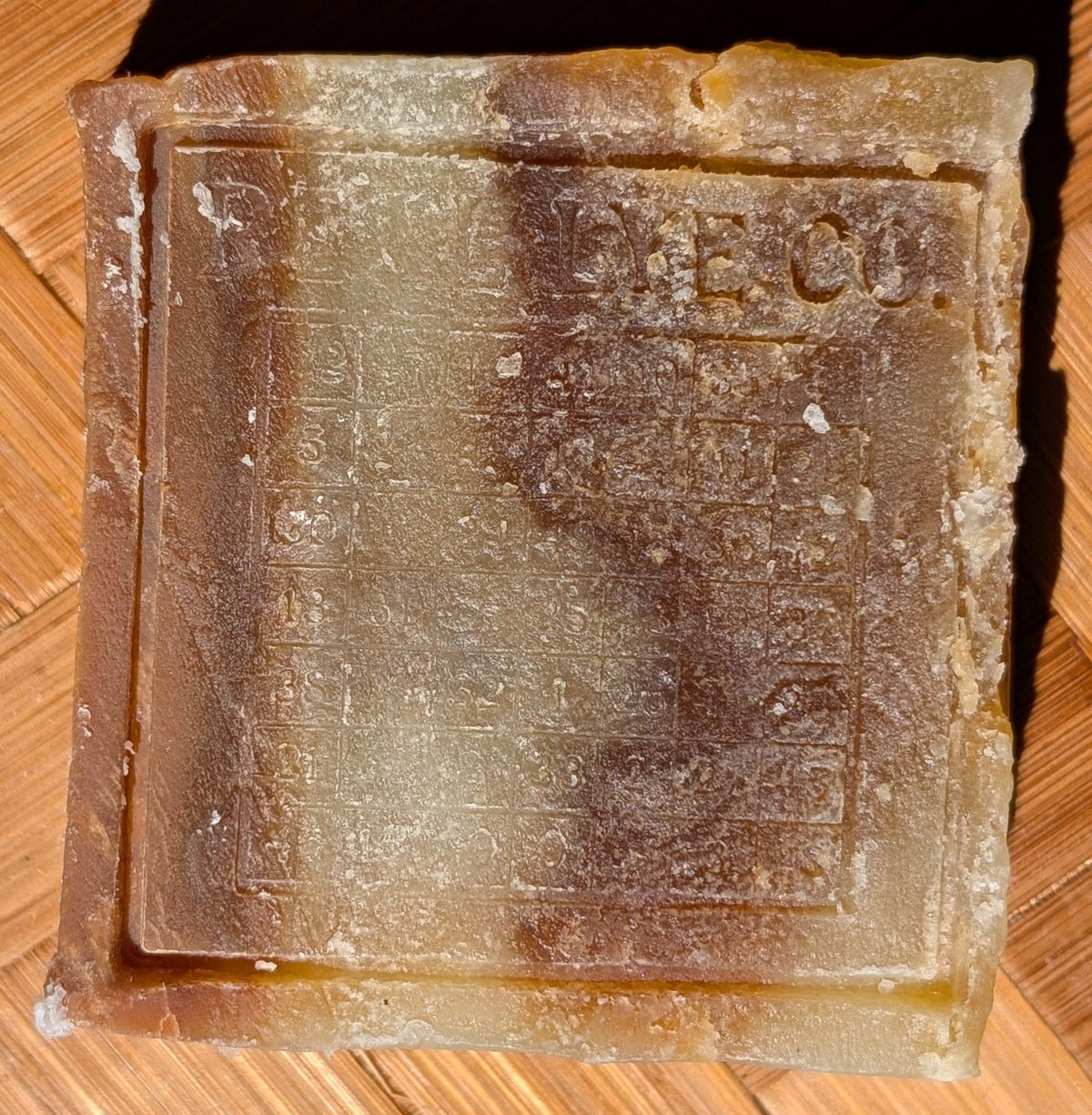 A square, rustic bar of soap with a raised border, resting on a woven bamboo mat under direct sunlight.