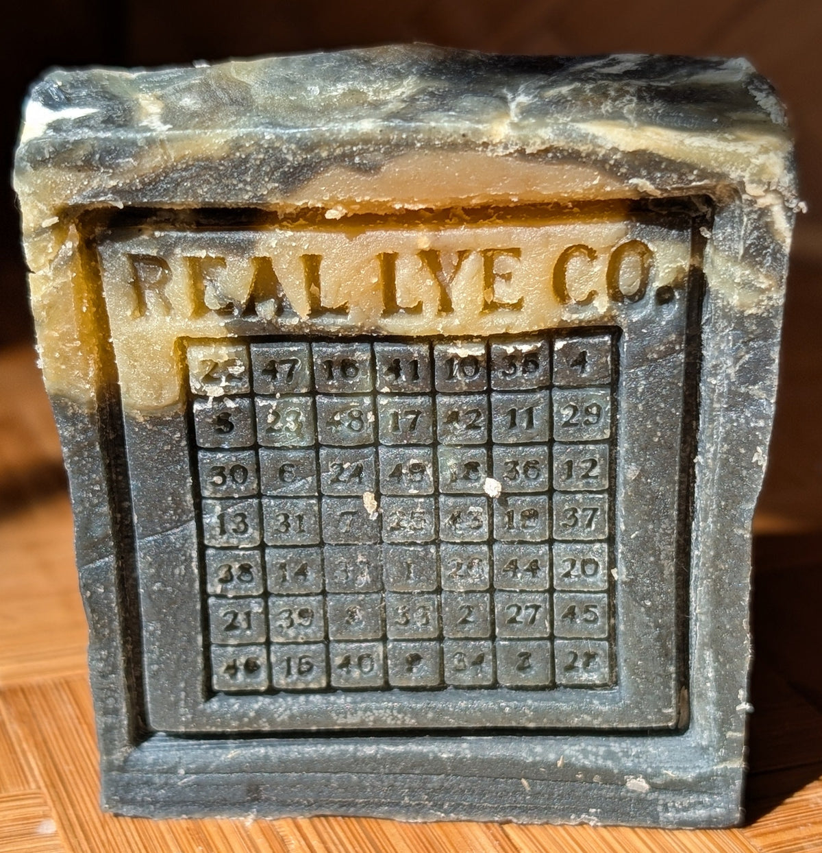 A vintage stone SOAP BAR featuring the embossed text 'REAL LYE CO.' and a grid of letterpress characters, photographed on a wooden parquet surface.