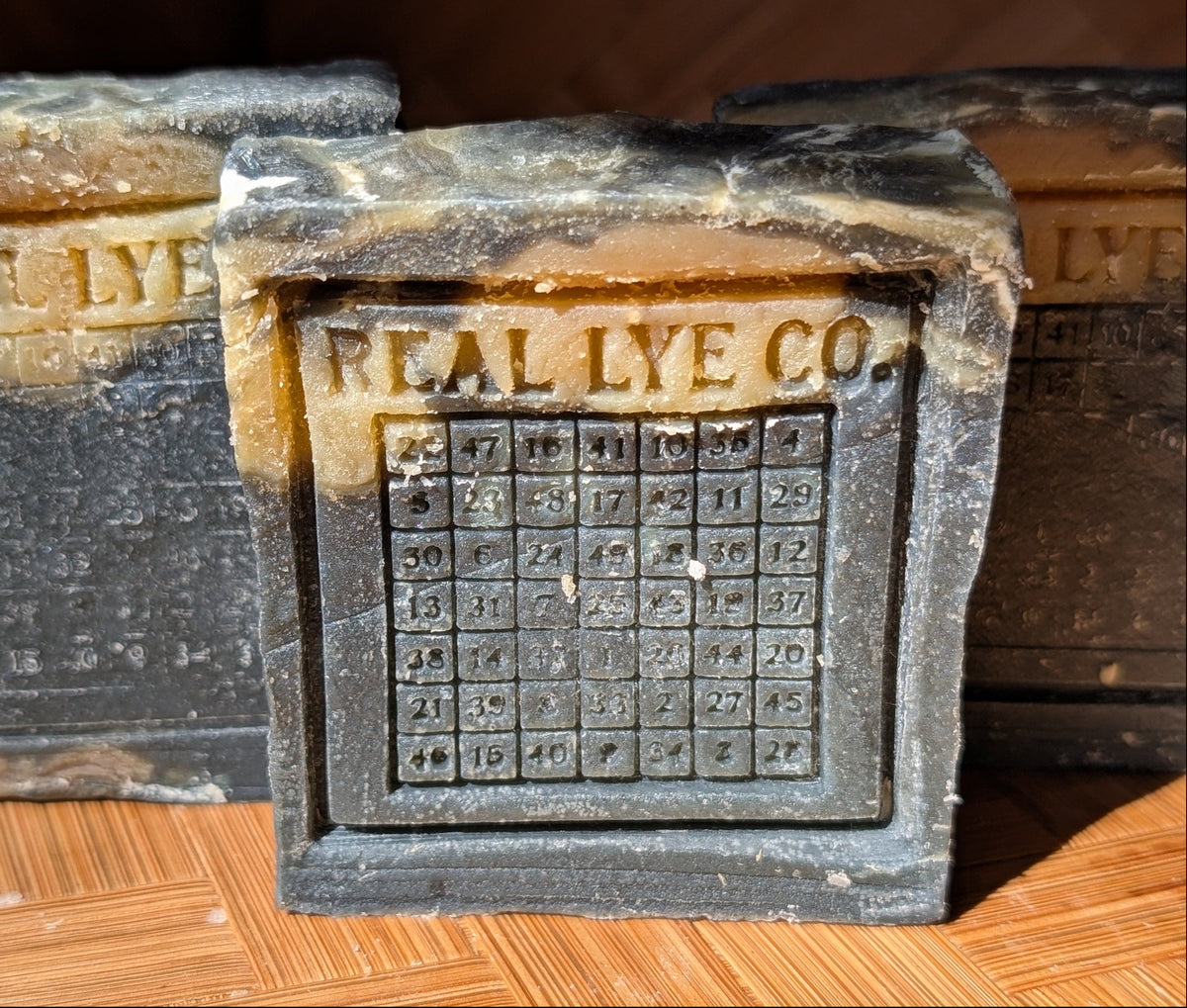 A close-up of a vintage-style novelty soap bar designed to look like a lye box, with 'REAL LYE CO.' embossed on the front above a grid of numbers, set on a wooden parquet floor.