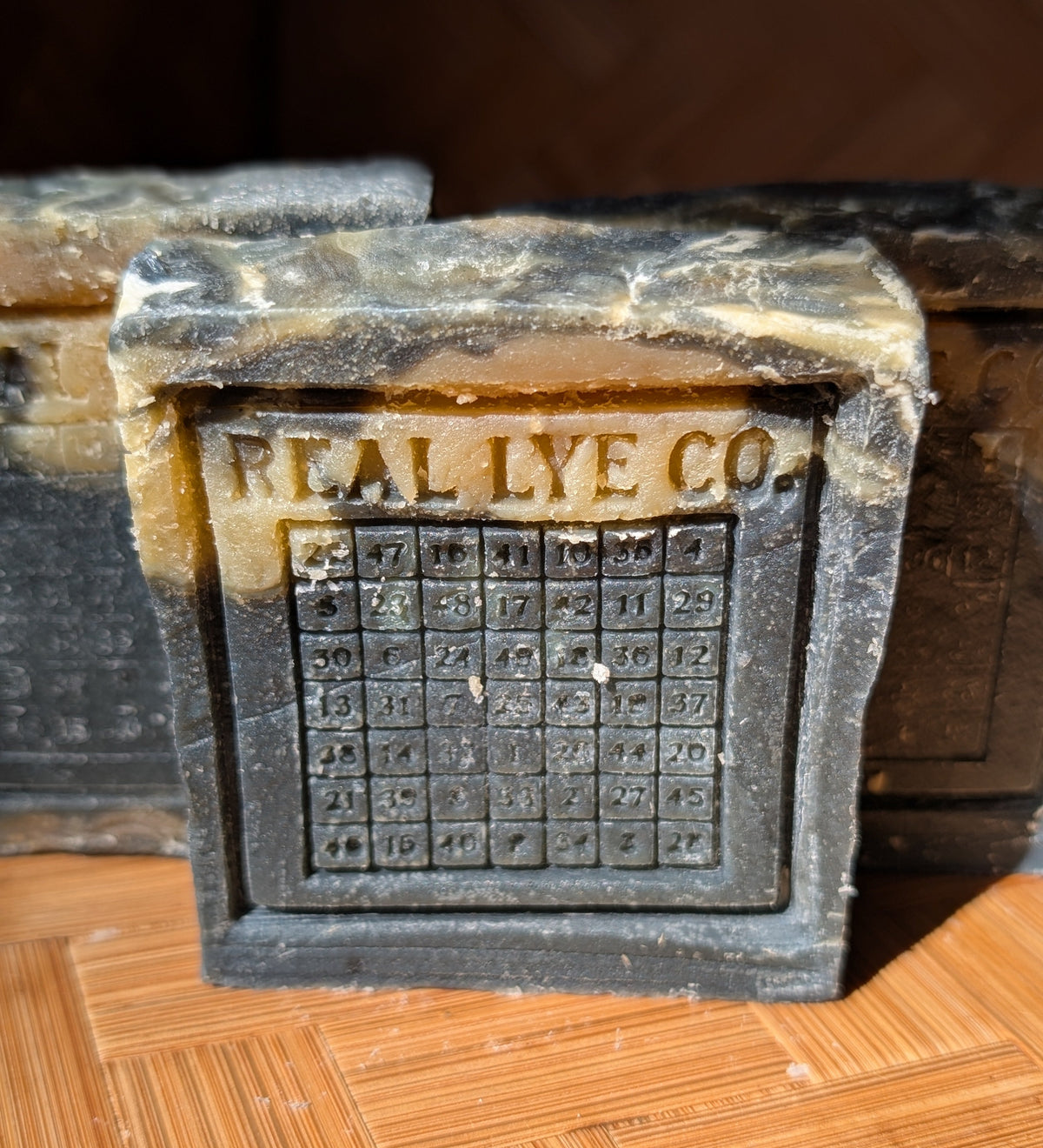 A close-up of a rectangular dark gray and tan stone-like plaque or block with the words 'REAL LYE CO.' and a grid of numbers, resting on a wood-patterned surface.