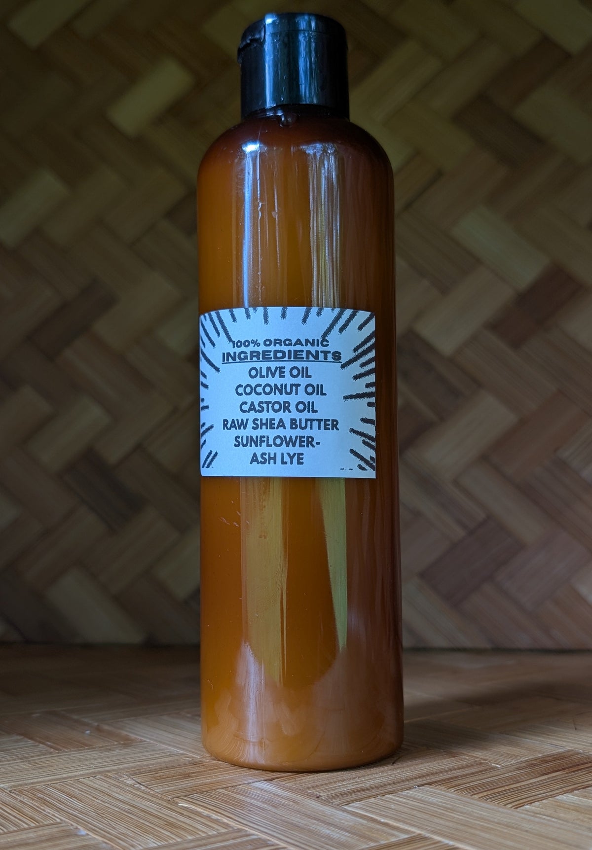An amber plastic bottle of organic olive oil and coconut oil hair conditioner with a black cap, standing on a wooden surface against a woven wicker background.