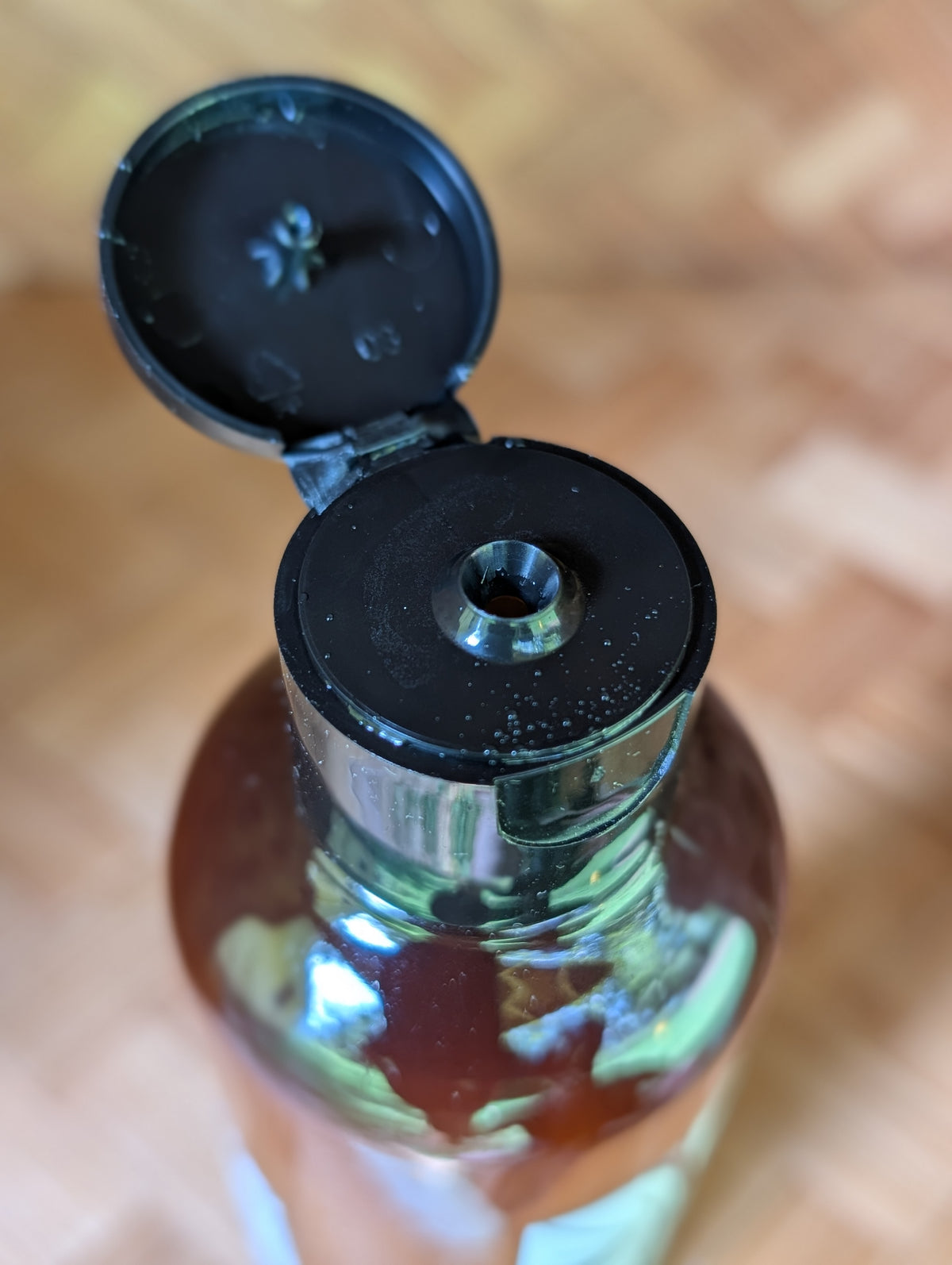 Close-up of a clear bottle with amber liquid and a black flip-top cap open, set against a blurred wooden background.