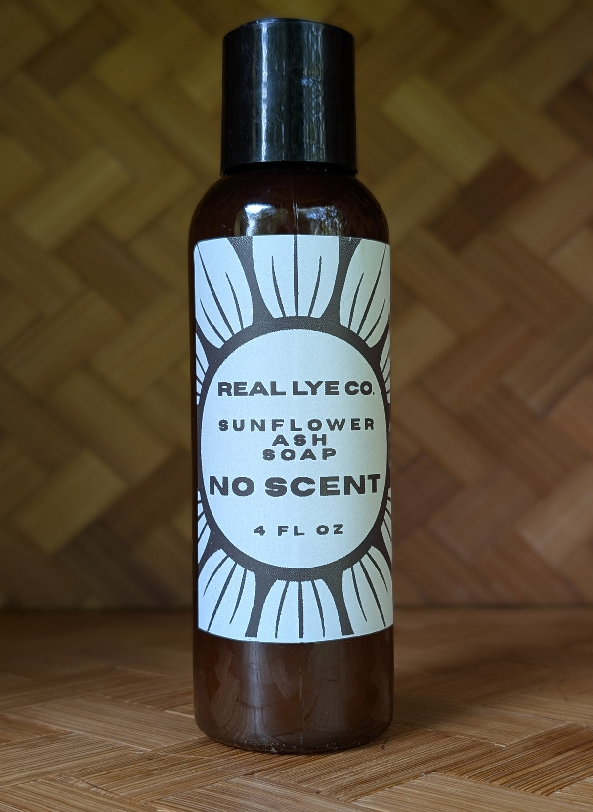 A 4 fl oz bottle of Real Lye Co Sunflower Ash Soap No Scent, featuring a dark amber-brown bottle, black cap, and a white label with a black sunburst design, set against a woven wood background.