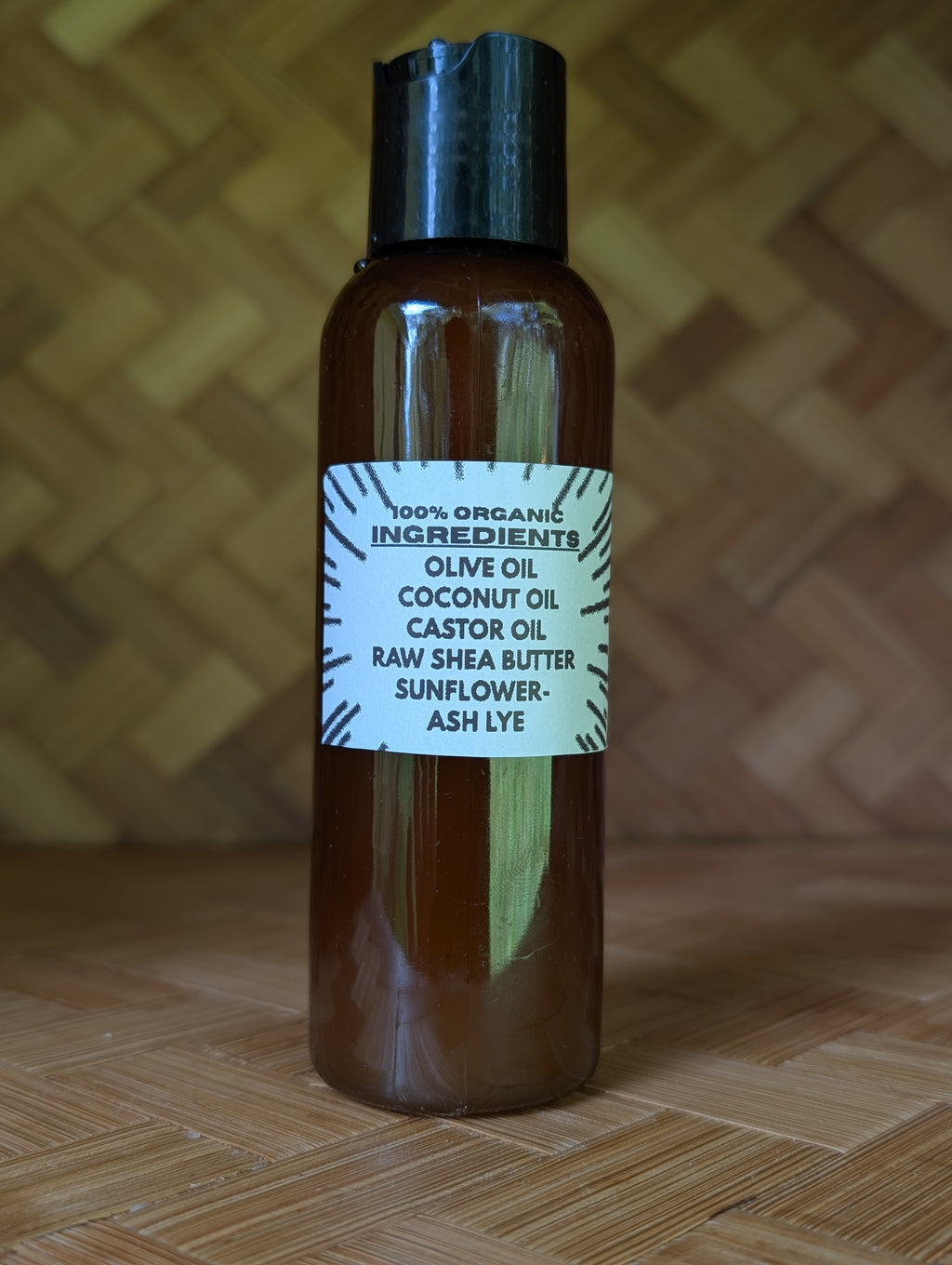 An amber glass bottle with a black cap and a white label listing ingredients such as olive oil, coconut oil, castor oil, raw shea butter, sunflower, and ash lye, set against a woven wood background.