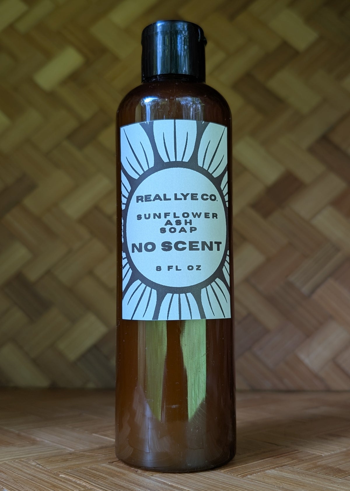 A 8 fl oz amber glass bottle of Real Lye Co Sunflower Ash Soap, No Scent, with a black cap and a white label, set against a woven wood background.