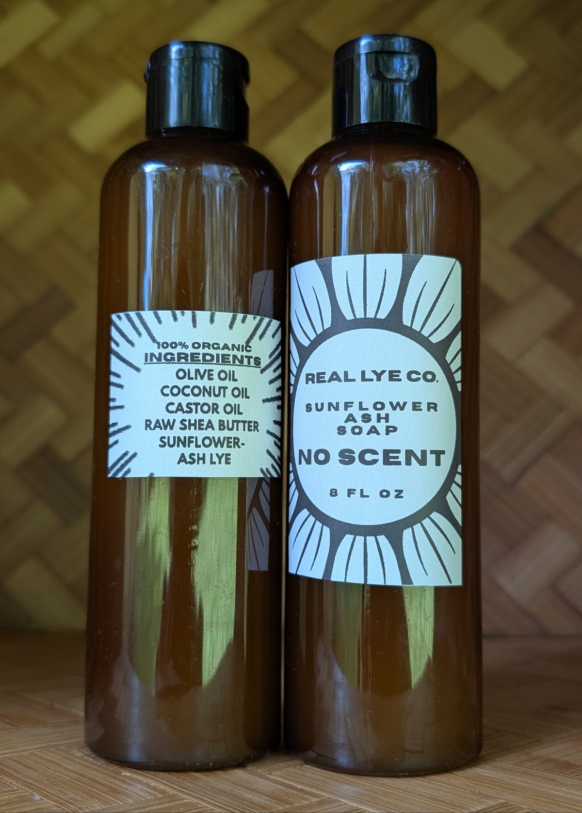 Two amber plastic bottles of bath products standing side by side on a wooden surface against a woven wood background. The left bottle has a white label reading '100% Organic Ingredients Olive Oil Coconut Oil Castor Oil Raw Shea Butter Sunflower-Ash Lye' and the right bottle has a white label reading 'REAL LYE CO SUNFLOWER SOAP NO SCENT 9 FL OZ'.