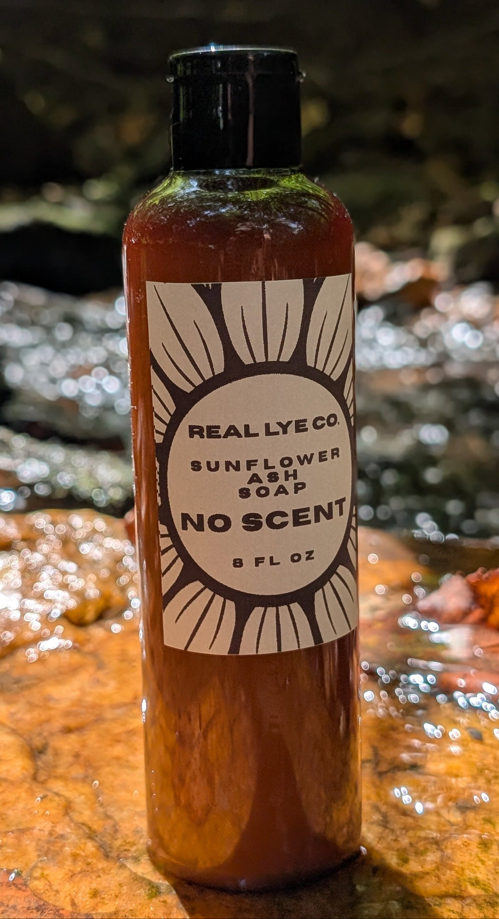 A bottle of Really Lye Co Sunflower Soap No Scent, 8 fl oz, standing on a wet rock in a stream. The bottle is amber-brown with a black cap and a white label featuring a sunflower design.