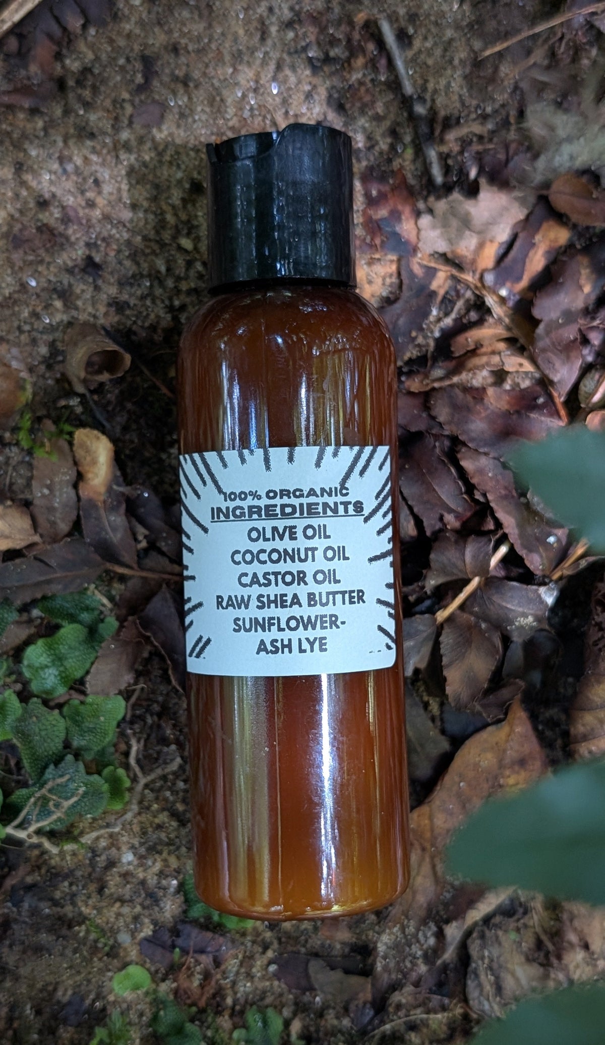 An amber glass bottle of hair oil with a black cap and a white label, lying on a bed of dry leaves and green moss outdoors.