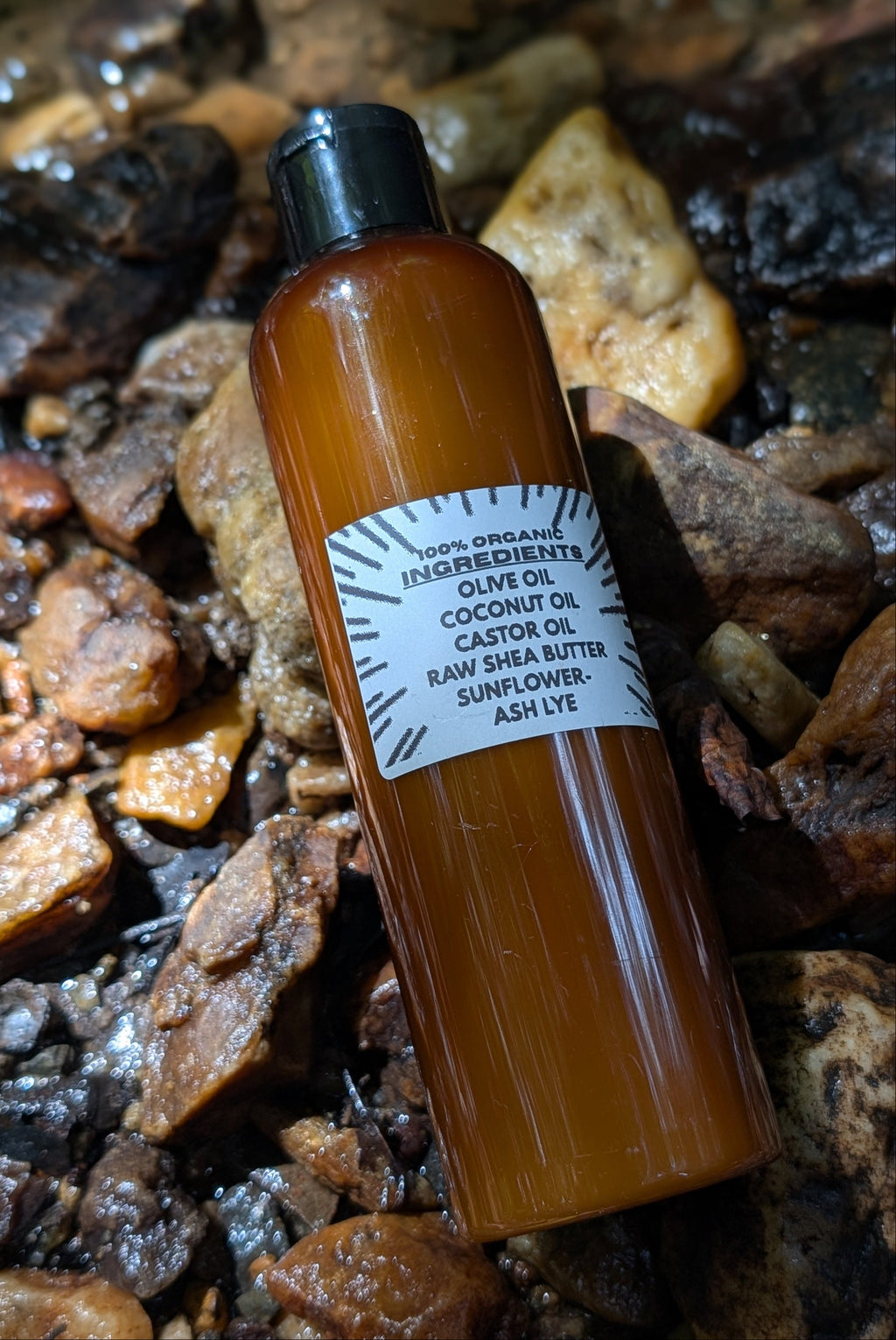 An amber glass bottle of raw unrefined olive oil with a black cap and a white label, placed diagonally on a bed of dark brown and tan stones.