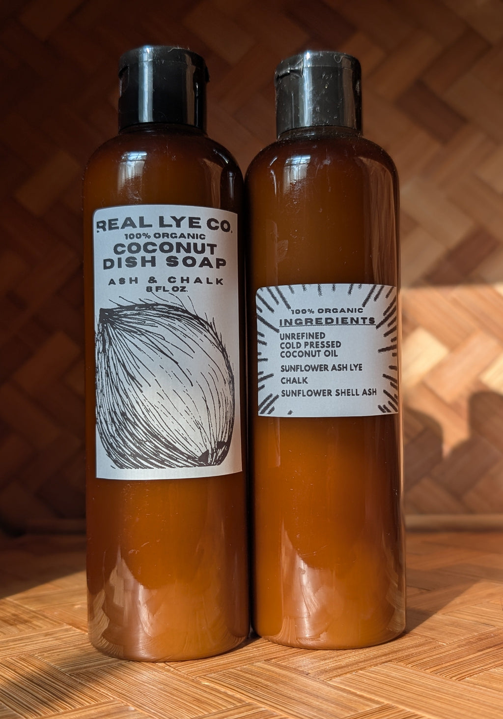 Two amber plastic bottles of Real Lye Co. coconut dish soap and coconut oil with black caps, standing side-by-side on a wooden surface against a wooden background.
