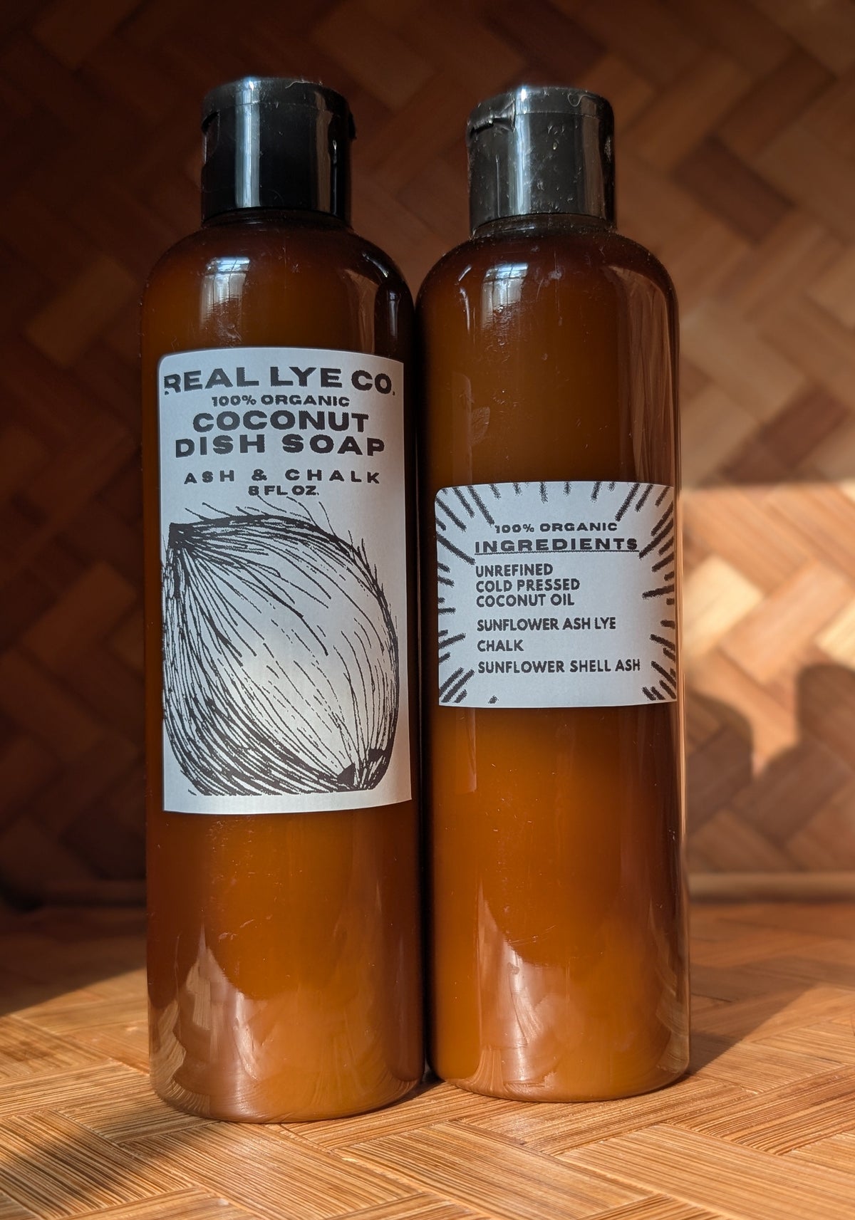 Two amber plastic bottles of Real Lye Co. coconut dish soap and coconut oil with black caps, standing side-by-side on a wooden surface against a wooden background.