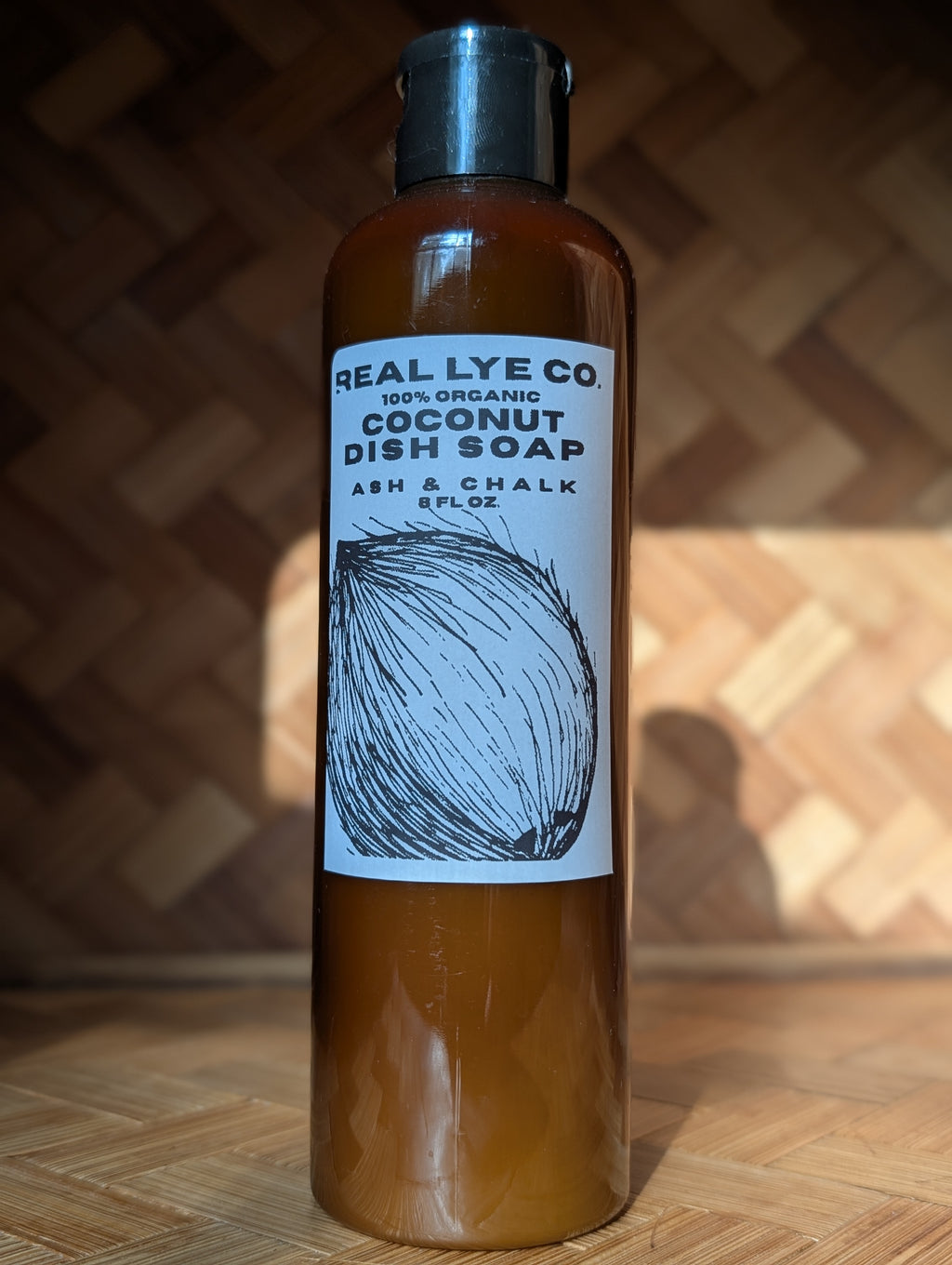 A bottle of Real Lye Co. Coconut Dish Soap standing upright on a wooden surface against a wooden herringbone-patterned background.