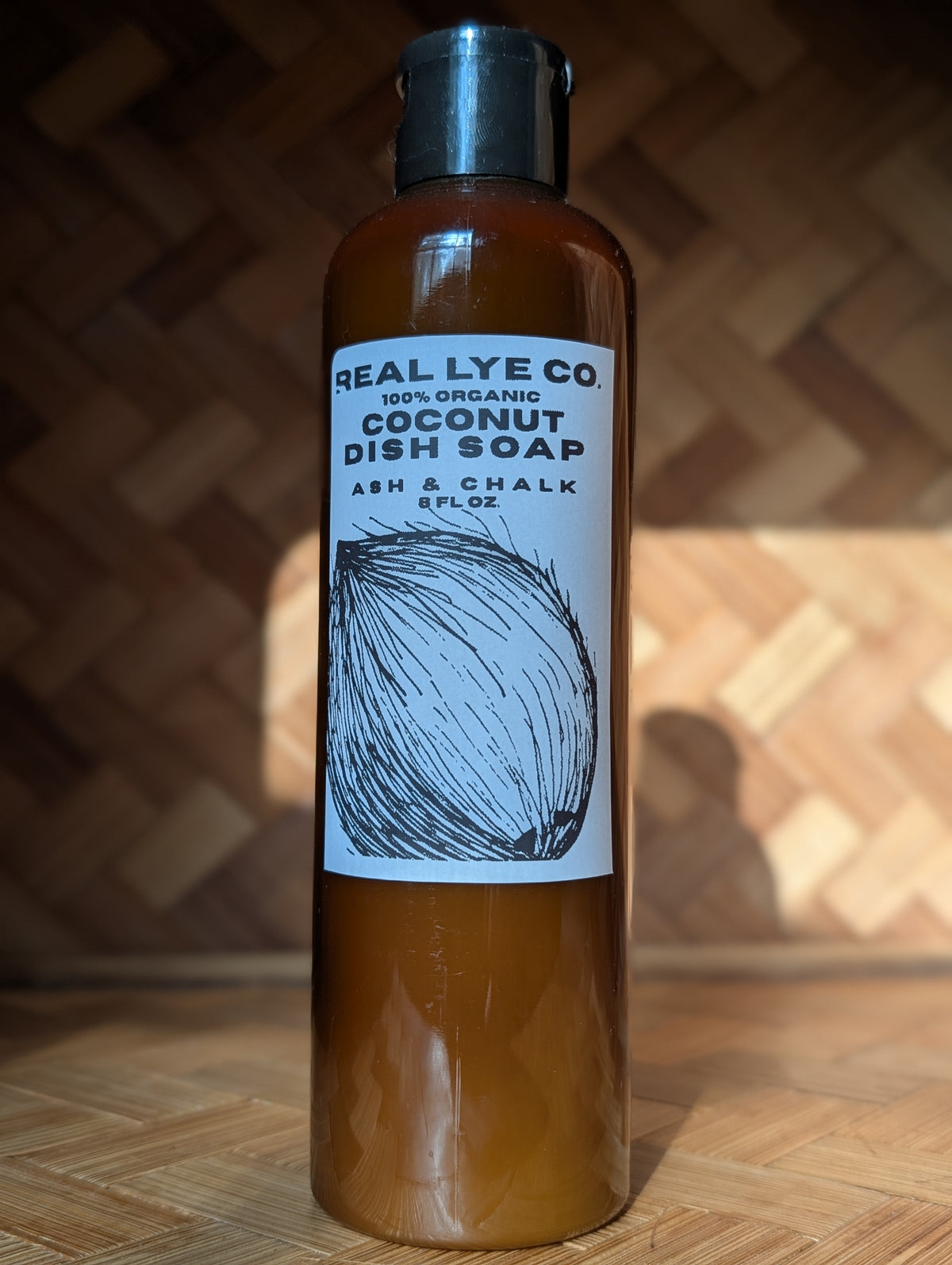 A bottle of Real Lye Co. Coconut Dish Soap standing upright on a wooden surface against a wooden herringbone-patterned background.