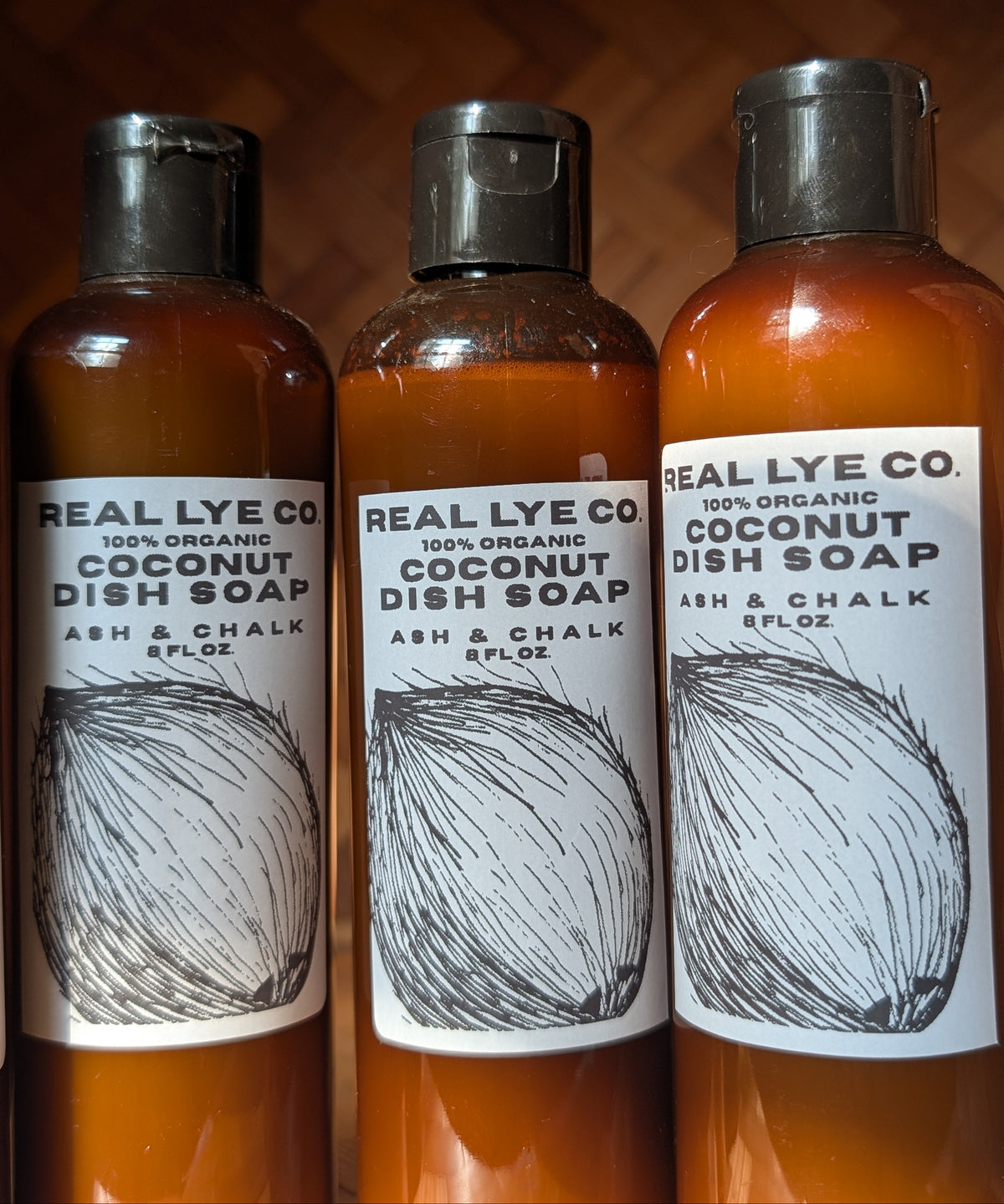 Three amber plastic bottles of Real Lye Co. Organic Coconut Dish Soap with black caps and white labels, arranged side-by-side against a dark brown background.