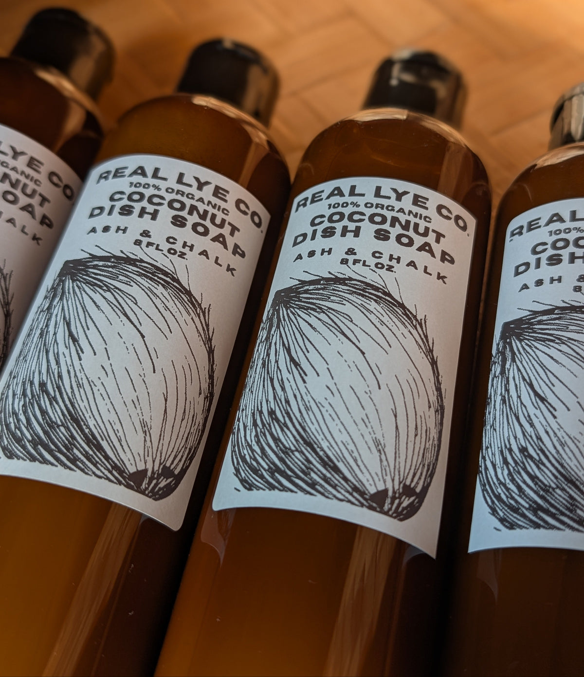 Four amber plastic bottles of Real Lye Co Coconut Dish Soap lined up on a wooden surface, showing white labels with black text and a coconut illustration.