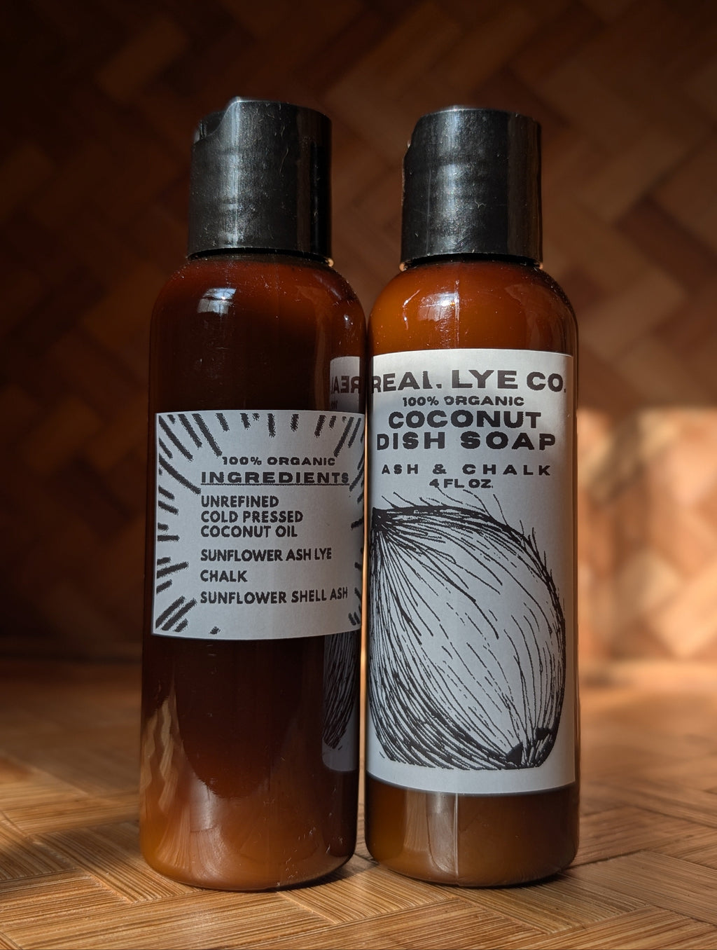 Two amber plastic bottles of dish soap and shampoo from Ash Real Lye Co standing side by side on a wooden surface against a warm wood-paneled background. The left bottle has a black cap and a white label reading '100% Organic Ingredients Sunflower Ash Lye Chalk Sunflower Shell Ash.