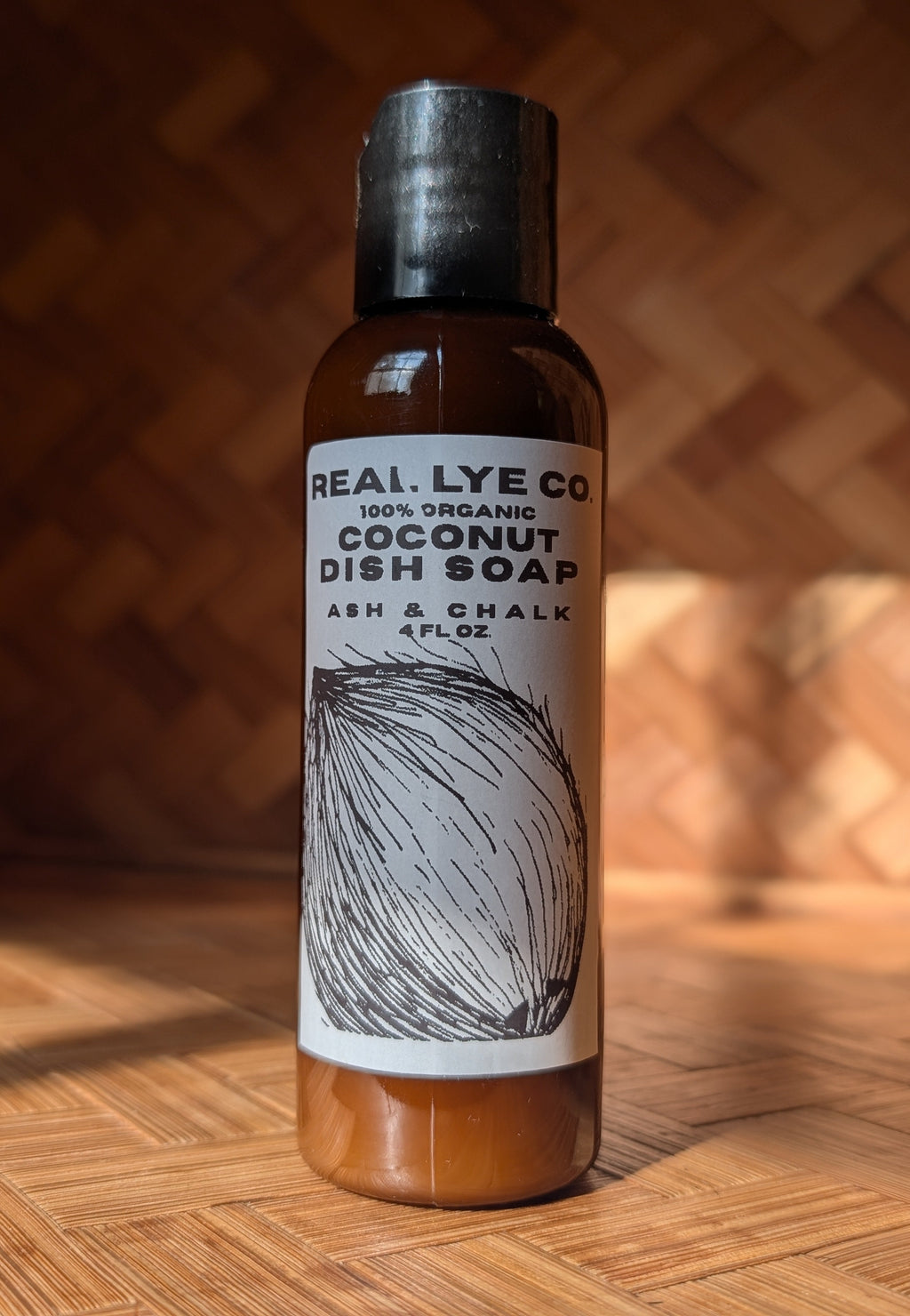 A bottle of Real Lye Co Coconut Dish Soap stands on a wooden surface against a wooden background. The bottle is amber brown with a black cap and a white label featuring a black line drawing of a coconut.