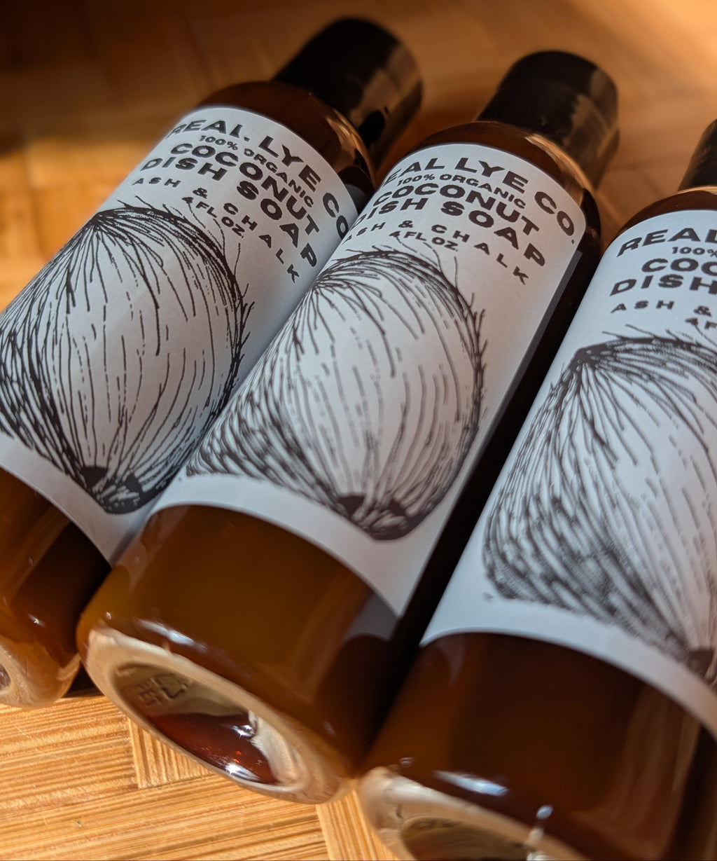 Three bottles of Real Lye Co. Organic Coconut Dish Soap arranged diagonally on a wooden surface, showing the brown soap liquid inside and white labels with black text and coconut illustrations.