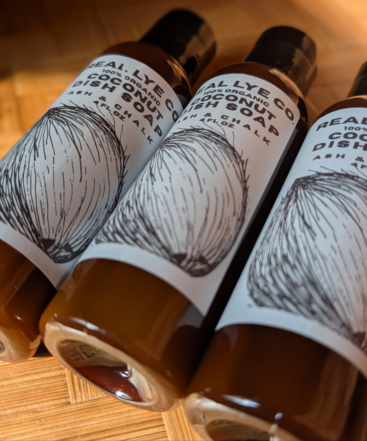 Three bottles of Real Lye Co. Organic Coconut Dish Soap arranged diagonally on a wooden surface, showing the brown soap liquid inside and white labels with black text and coconut illustrations.
