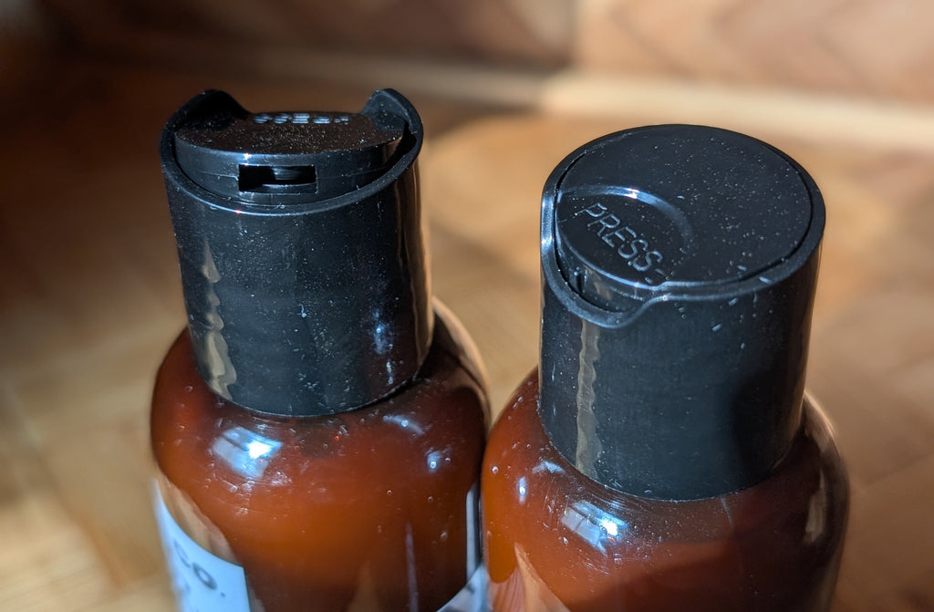 Two amber plastic bottles with black flip-top caps, shown close up on a wooden surface with a blurred woven background.