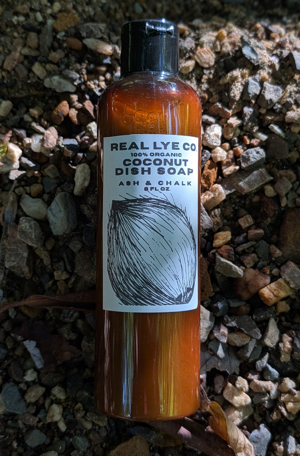 A bottle of Real Lye Co Coconut Dish Soap stands upright on a bed of small gray and brown gravel with scattered dry leaves. The bottle is amber-brown with a black cap and a white label featuring a black coconut illustration.