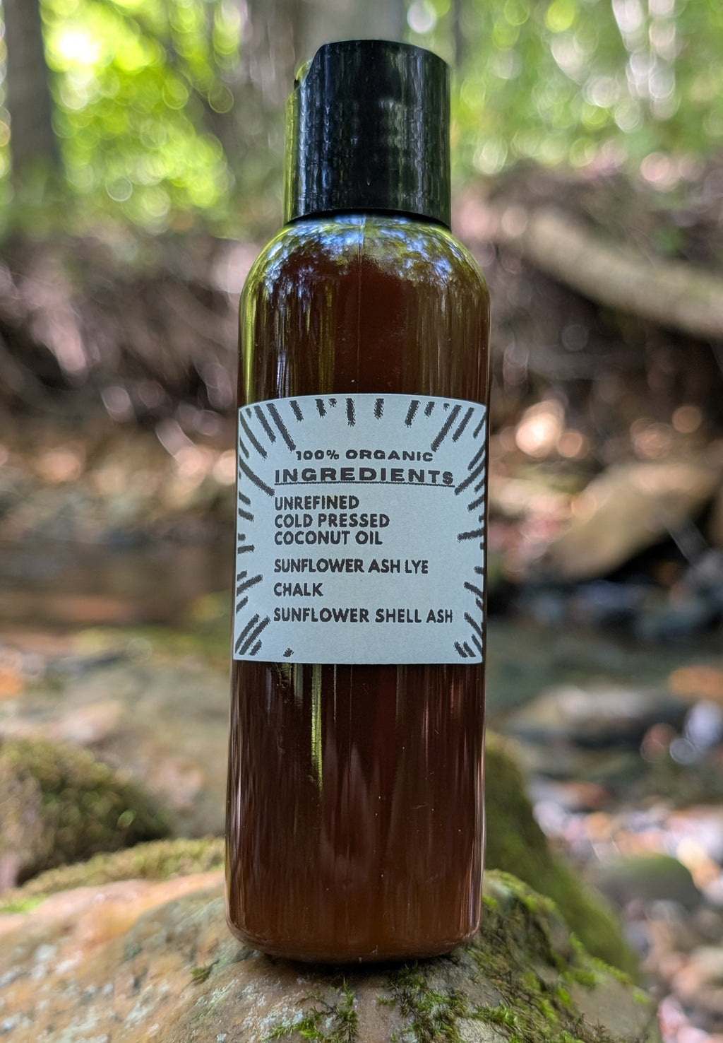A bottle of cold pressed coconut oil with a black cap and white label, standing on a mossy log in a forest setting.
