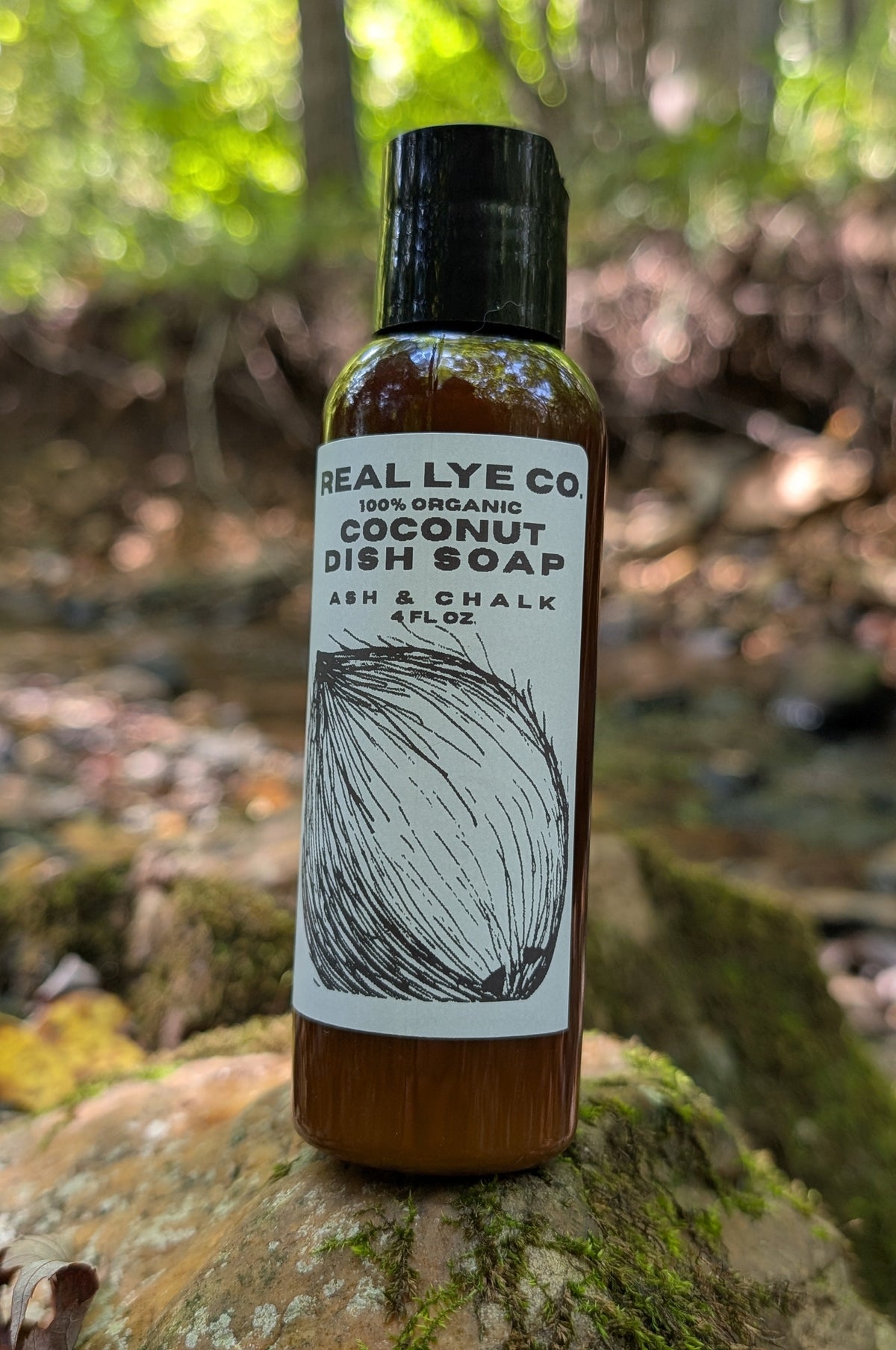 A bottle of Real Lye Co Coconut Dish Soap standing on a mossy log in a forest setting. The bottle is amber brown with a black cap and a white label featuring a black line drawing of a coconut.